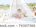 A little cute girl with red curly hair in summer, on the seashore in an airy lace bodysuit and a floral headband in a lace tent, devoried with marine details, shells and cotton. 1st BIRTHDAY.  74307366