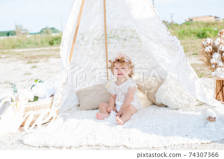 A little cute girl with red curly hair in summer, on the seashore in an airy lace bodysuit and a floral headband in a lace tent, devoried with marine details, shells and cotton. 1st BIRTHDAY.  74307366
