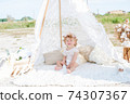 A little cute girl with red curly hair in summer, on the seashore in an airy lace bodysuit and a floral headband in a lace tent, devoried with marine details, shells and cotton. 1st BIRTHDAY.  74307367