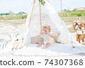 A little cute girl with red curly hair in summer, on the seashore in an airy lace bodysuit and a floral headband in a lace tent, devoried with marine details, shells and cotton. 1st BIRTHDAY.  74307368