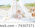 A little cute girl with red curly hair in summer, on the seashore in an airy lace bodysuit and a floral headband in a lace tent, devoried with marine details, shells and cotton. 1st BIRTHDAY.  74307371