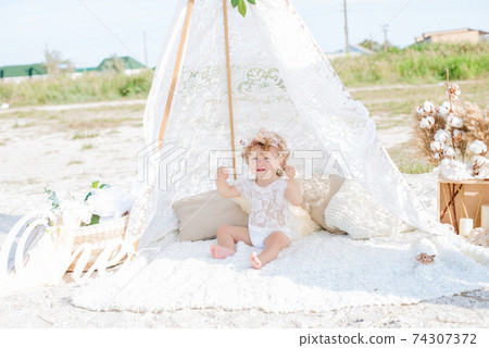 A little cute girl with red curly hair in summer, on the seashore in an airy lace bodysuit and a floral headband in a lace tent, devoried with marine details, shells and cotton. 1st BIRTHDAY.  74307372