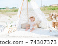 A little cute girl with red curly hair in summer, on the seashore in an airy lace bodysuit and a floral headband in a lace tent, devoried with marine details, shells and cotton. 1st BIRTHDAY.  74307373