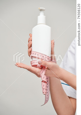 Portrait of beauty specialist standing against grey background and holding in her hand bottle of body lotion and measure tape 74308120