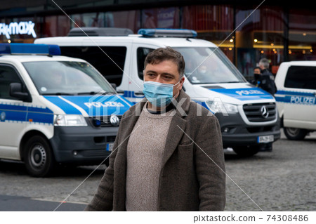 Outdoor portrait of a man protecting his face with a medical or surgical mask. On the background, out of focused police cars Outdoor portrait of a man protecting his face with a medical or surgical mask. On the background, out of focused police cars 74308486