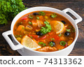 Fish soup with salmon, tilapia, red pepper, onion, carrot, lemon, green and black olives. White casserole on wooden rustic table, close-up 74313362