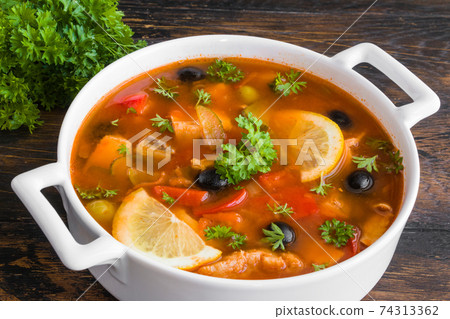 Fish soup with salmon, tilapia, red pepper, onion, carrot, lemon, green and black olives. White casserole on wooden rustic table, close-up Fish soup with salmon, tilapia, red pepper, onion, carrot, lemon, green and black olives. White casserole on wooden rustic table, close-up 74313362