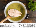 Fish broth in casserole, piece of salmon on spoon, green parsley, top view 74313369