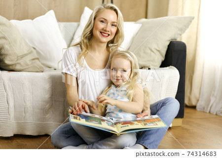 Happy family. Blonde young mother reading a book to her cute daughter while sitting at wooden floor. Motherhood concept 74314683