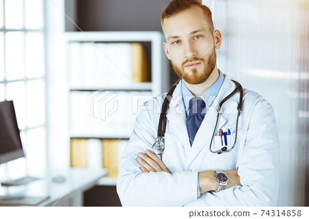 Red-bearded doctor standing straight in sunny clinic near his working place. Portrait of physician. Medicine concept Red-bearded doctor standing straight in sunny clinic near his working place. Portrait of physician. Medicine concept 74314858