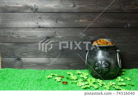 Cast iron pot with four-petal lucky shamrock leaf, full of leprechaun gold treasure, shamrock leaves and coins on green grass and dark wooden planks background. St. Patricks Day banner, poster, flyer 74316040