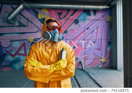 Graffiti painter in respirator mask standing near the wall with his paintings Graffiti painter in respirator mask standing near the wall with his paintings 74318875