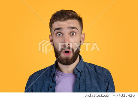 Unbelievable news. Portrait of bearded young guy opening mouth and eyes in shock, looking at camera on orange background Unbelievable news. Portrait of bearded young guy opening mouth and eyes in shock, looking at camera on orange background 74323420
