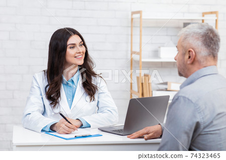 Beautiful doctor in white medical coat listening to old patient and making notes in tablet Beautiful doctor in white medical coat listening to old patient and making notes in tablet 74323465