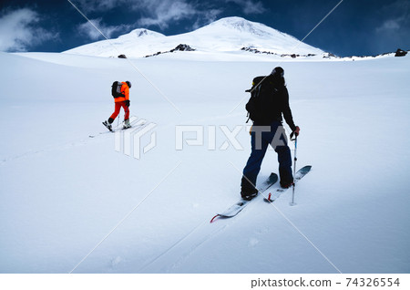 Two freeride skiers climb up the fresh snow in a ski tour. The summit of Mount Elbrus and the blue sky on the background Two freeride skiers climb up the fresh snow in a ski tour. The summit of Mount Elbrus and the blue sky on the background 74326554