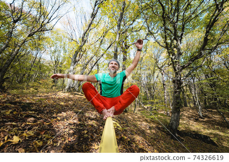 A bearded man in age balances while sitting on a taut slackline in the autumn forest. Outdoor Leisure A bearded man in age balances while sitting on a taut slackline in the autumn forest. Outdoor Leisure 74326619