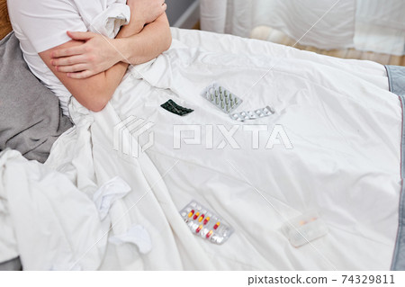 ill young caucasian man sitting on bed having high fever, surrounded by medications ill young caucasian man sitting on bed having high fever, surrounded by medications 74329811