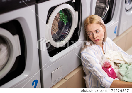 Laundry interior, young caucasian woman sitting near washing machines and looking up sadly 74329897
