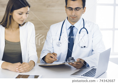 Doctor and patient discussing medical exam results while sitting at the desk in clinic. Male physician using clipboard for filling up medication history record of young woman. Data in medicine Doctor and patient discussing medical exam results while sitting at the desk in clinic. Male physician using clipboard for filling up medication history record of young woman. Data in medicine 74342687