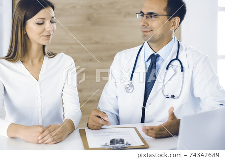 Doctor and patient discussing medical exam results while sitting at the desk in clinic. Male physician using clipboard for filling up medication history record of young woman. Data in medicine Doctor and patient discussing medical exam results while sitting at the desk in clinic. Male physician using clipboard for filling up medication history record of young woman. Data in medicine 74342689