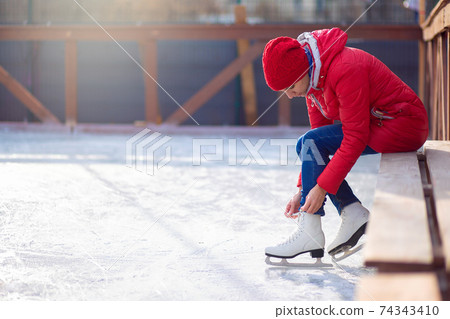 A girl in a red jacket sits on a bench on an open ice rink and laces figure skates. Winter sport 74343410