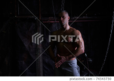 Young shirtless muscular man standing among metal chains, looking at camera, copy space 74343730