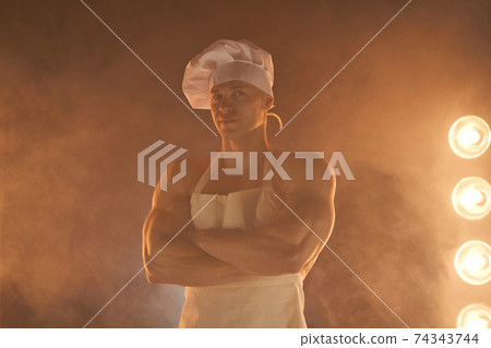 Portrait of muscular chef wearing white apron and chef hat, posing with folded arms on smoky background 74343744