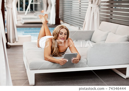 A young woman in a bikini relaxes with a tablet by the pool on a sunny day. Online business, freelance 74346444