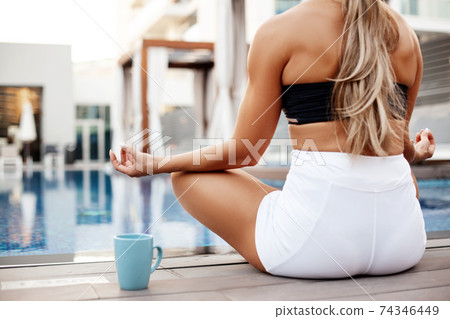 A young girl is sitting by the pool in the lotus position, meditating and doing yoga exercises. Yoga, fitness, healthy lifestyle. Rear view A young girl is sitting by the pool in the lotus position, meditating and doing yoga exercises. Yoga, fitness, healthy lifestyle. Rear view 74346449