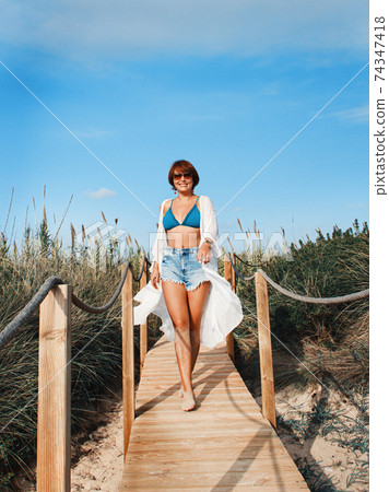 Young woman in cover up and jeans shorts walking between dunes. Young woman in cover up and jeans shorts walking between dunes. 74347418