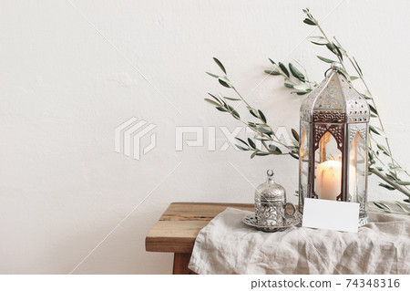 Ramadan Kareem greeting card, invitation mockup. Silver lantern with burning candle and silver cup with tea or coffee. Green olive tree branches on old wooden table background. Muslim Iftar dinner. Ramadan Kareem greeting card, invitation mockup. Silver lantern with burning candle and silver cup with tea or coffee. Green olive tree branches on old wooden table background. Muslim Iftar dinner. 74348316