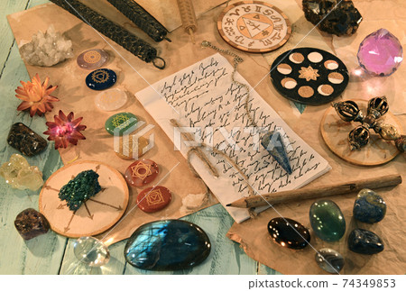 Crystal stones with chakras, candles, ritual objects and paper with lettering and text on the table. Esoteric, gothic and occult background. No foreign language, all symbols are fantasy 74349853