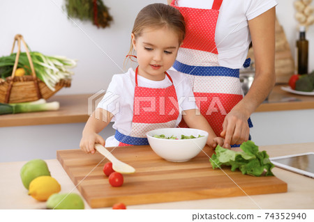 Happy family in the kitchen. Mother and child daughter cooking tasty breakfest of fresh salad. Little helper slicing and mix tomatos and greenery Happy family in the kitchen. Mother and child daughter cooking tasty breakfest of fresh salad. Little helper slicing and mix tomatos and greenery 74352940