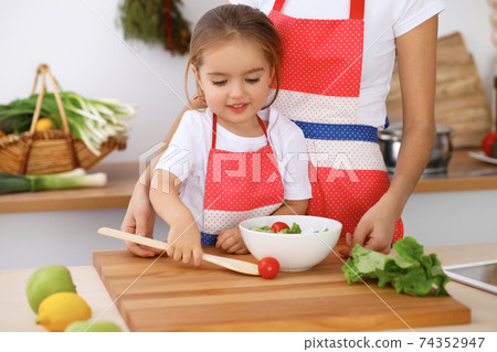 Happy family in the kitchen. Mother and child daughter cooking tasty breakfest of fresh salad. Little helper slicing and mix tomatos and greenery 74352947