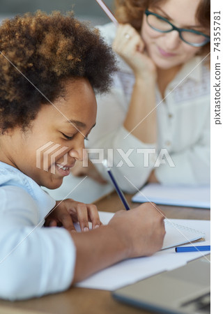 Close up shot of enthusiastic mixed race teen schoolgirl writing with pencil during a lesson with her female teacher at home Close up shot of enthusiastic mixed race teen schoolgirl writing with pencil during a lesson with her female teacher at home 74355781