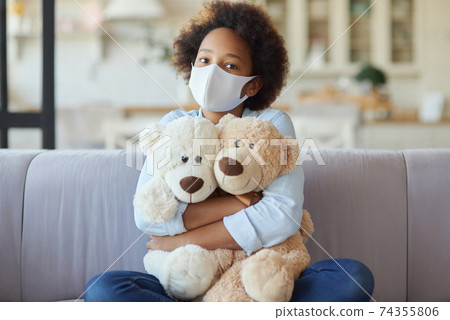 Portrait of cute mixed race teen girl in casual clothes wearing protective face mask, looking at camera, hugging her toys while sitting on a sofa at home Portrait of cute mixed race teen girl in casual clothes wearing protective face mask, looking at camera, hugging her toys while sitting on a sofa at home 74355806