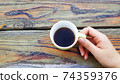 A photo of a hand holding a mug with coffee on a wooden table 74359376