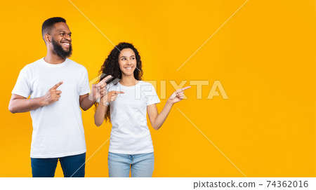 Multiracial Couple Pointing Fingers Aside Over Yellow Studio Background, Panorama Multiracial Couple Pointing Fingers Aside Over Yellow Studio Background, Panorama 74362016