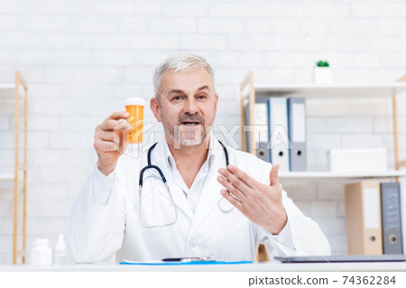 Smiling mature male physician in white medical uniform sit at desk in hospital, medicine, healthcare 74362284