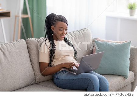 Joyful african lady sitting on couch at home, using laptop 74362422