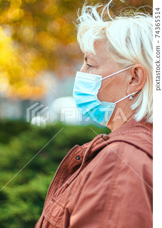 Caucasian woman 50 years old in a protective medical mask looks away in an autumn park. Safety in public place during coronavirus outbreak. Caucasian woman 50 years old in a protective medical mask looks away in an autumn park. Safety in public place during coronavirus outbreak. 74365514