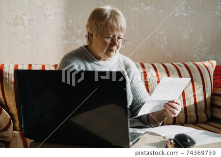 Senior woman using laptop and holding documents at home. Focused mature aged female using computer working with finance bank loan or mortgage documents online Senior woman using laptop and holding documents at home. Focused mature aged female using computer working with finance bank loan or mortgage documents online 74369739