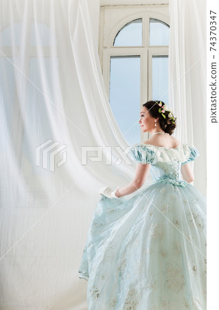 Beautiful elegant dark haired woman in historic dress on background with window 74370347