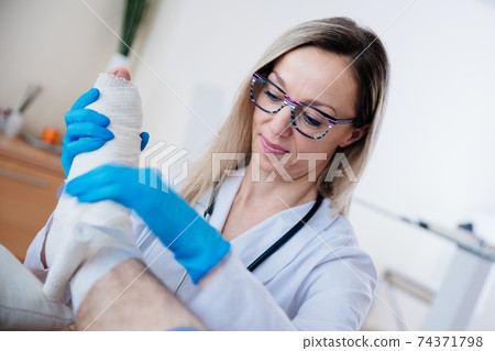 Smiling professional nurse with glasses is bandaging the leg. Treatment of broken bones. 74371798
