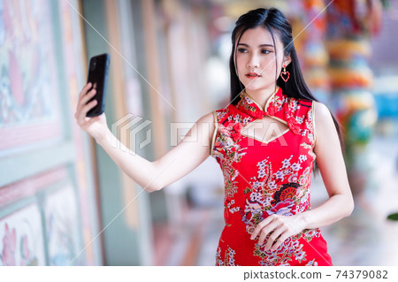 Portrait beautiful smiles Asian young woman wearing red traditional Chinese cheongsam decoration taking a selfie with smartphone for Chinese New Year Festival at Chinese shrine 74379082