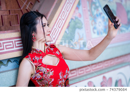 Portrait beautiful smiles Asian young woman wearing red traditional Chinese cheongsam decoration taking a selfie with smartphone for Chinese New Year Festival at Chinese shrine 74379084