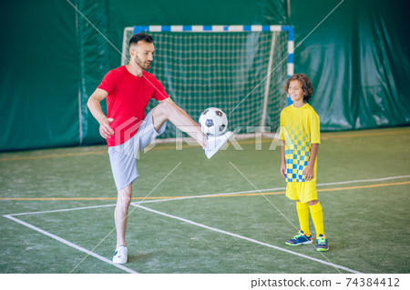 Man in a red tshirt kicking a ball, a boy watching him 74384412