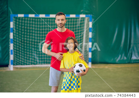 PE teacher in red tshirt standing near his pupil and holding a ball PE teacher in red tshirt standing near his pupil and holding a ball 74384443