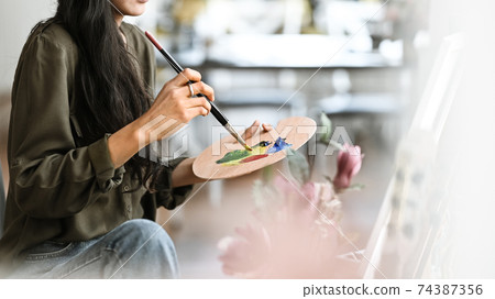 Cropped image of young beautiful artist girl sitting in front the drawing canvas while painting an oil color at the modern studio. Beautiful artist and gallery studio concept. Cropped image of young beautiful artist girl sitting in front the drawing canvas while painting an oil color at the modern studio. Beautiful artist and gallery studio concept. 74387356