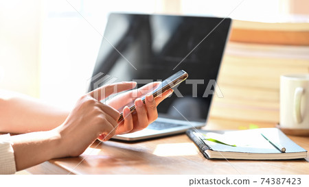 Cropped image of woman's hands holding/using smartphone at the modern wooden desk. Cropped image of woman's hands holding/using smartphone at the modern wooden desk. 74387423